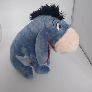 Puede incluir: Un peluche de Eeyore, el burro de Winnie the Pooh. El juguete es azul con una melena negra, un hocico rosa y el interior de la oreja rojo. Está sobre una superficie blanca.