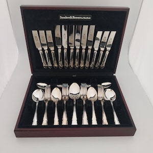 May include: A boxed set of silver cutlery, including knives, forks, and spoons. The silverware is arranged neatly within a dark wooden box lined with black velvet. The box is branded with the text "Sanders & Bowers of Sheffield".