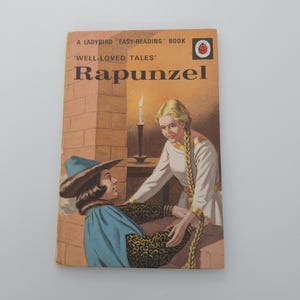 Pode incluir: Um livro vintage Ladybird Easy-Reading intitulado "Rapunzel". A capa apresenta uma ilustração de Rapunzel com longas tranças loiras e um homem com capa e chapéu azuis. O livro tem um logotipo Ladybird vermelho e branco.