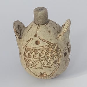 May include: A beige ceramic bottle with a cylindrical stopper. The bottle has two small ear-like protrusions and is decorated with etched geometric patterns, including triangles and circles. The overall design suggests a handcrafted, artisanal aesthetic.