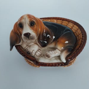 May include: A ceramic Basset Hound puppy figurine in a woven basket. The dog has brown and white markings, with long floppy ears and a sad expression. The basket is brown with a textured design.