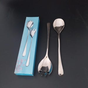 May include: A set of polished stainless steel serving utensils, including a spoon, a slotted spoon, and a fork-like serving utensil. The utensils are displayed next to a light blue box with a floral design.