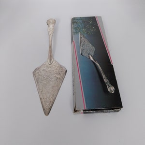 Puede incluir: Servidor de pastel de plata con un diseño ornamentado. El servidor está en su caja original, que es azul y blanca con las palabras "Cake Server" impresas en ella.