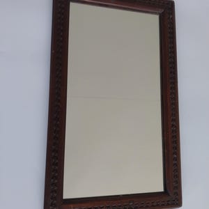 May include: Rectangular mirror with a dark brown, intricately carved wooden frame. The frame features a repeating geometric pattern along its edges. The mirror's surface reflects a neutral, off-white background.