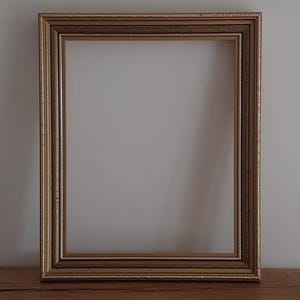 May include: A rectangular, gold-colored picture frame with a detailed, ornate design. The frame has a double border and sits on a wooden surface. The interior is empty, ready for a photo or artwork.