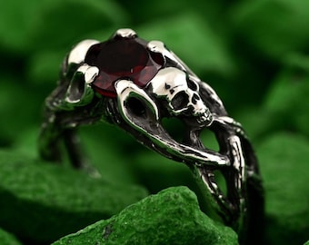 May include: A silver ring featuring a dark red gemstone, set within a design of skulls and intertwined branches. The ring is displayed against a backdrop of green, textured stones, creating a striking contrast.