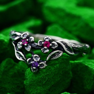 May include: A silver ring with a floral design featuring three gemstones. Two of the gemstones are pink and one is purple.