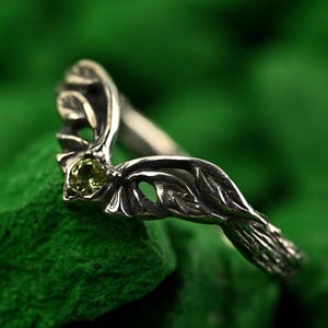 Könnte beinhalten: Ein silberfarbener Ring mit einem blattartigen Design und einem kleinen, runden, hellgrünen Edelstein. Der Ring ist detailliert und elegant. Er ist vor einem grünen Hintergrund platziert.