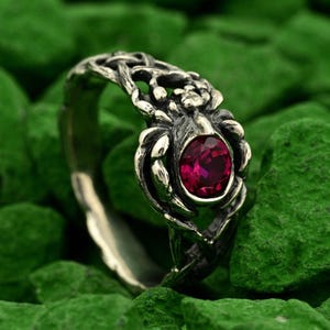 May include: A silver ring with an ornate design featuring a central oval-cut, deep red gemstone. The ring's band has a textured, interwoven pattern, and the gemstone is set within a detailed, stylized setting.