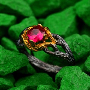 Peut inclure: Gros plan sur une bague ornée d'une grosse pierre précieuse rouge vif sertie dans un anneau en métal doré et noir. Le design de la bague est inspiré de la nature, avec une monture en forme de branches entrelacées.