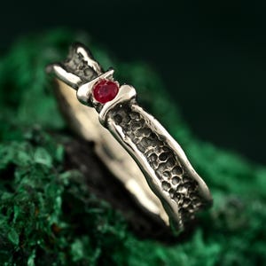 May include: A silver ring with a textured band and a small, round, red gemstone. The ring is set against a green background, highlighting the contrast between the metal and the gem.