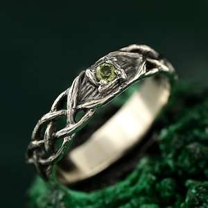 Puede incluir: Un anillo de plata con un diseño de nudo celta y una pequeña gema redonda de color verde claro. El anillo tiene un acabado texturizado y oxidado, y la gema está engastada en un engaste en forma de hoja. El anillo se muestra sobre una superficie verde texturizada.