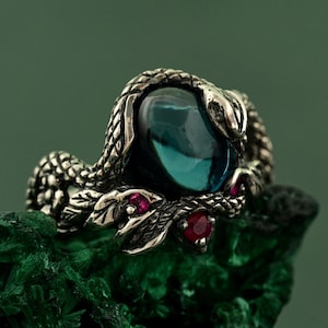 Op de afbeelding: Zilveren ring met een slangenmotief, gewikkeld rond een grote, teal-kleurige edelsteen. De kop van de slang rust boven de steen, met kleine rode edelstenen als accent. De ring staat tegen een groene achtergrond.