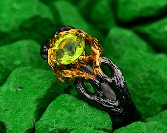 Op de afbeelding: Een close-up van een ring met een grote, ovale, lichtgroene edelsteen. De ring heeft een goudkleurige zetting rond de steen en een zwarte band met een ingewikkeld, getextureerd ontwerp. De ring is geplaatst tegen een achtergrond van heldergroene stenen.