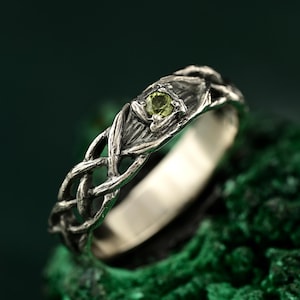 Könnte beinhalten: Ein silberner Ring mit einem keltischen Knotenmuster. Der Ring hat einen kleinen, runden, hellgrünen Edelstein in der Mitte. Das Band hat ein kompliziertes, ineinander verwobenes Muster. Der Ring wird auf einer strukturierten, dunkelgrünen Oberfläche präsentiert.