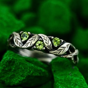 Può includere: Anello in argento con design a foglie e tre gemme rotonde verde chiaro. L'anello è incastonato su uno sfondo di rocce verdi, creando un'estetica ispirata alla natura. I dettagli intricati dell'anello sono evidenziati.