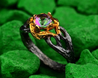 Op de afbeelding: Een close-up van een edelsteenring met een veelkleurige iriserende steen. De ring heeft een zwarte band met goudkleurige accenten rond de steen. De ring staat tegen een achtergrond van groene stenen.