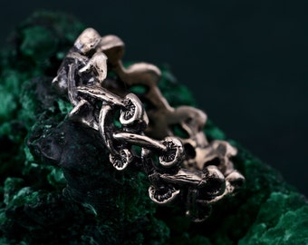 May include: A metallic ring featuring a continuous band design composed of multiple small, sculpted mushroom shapes. The ring has a dark, oxidized finish with visible texture on the caps and stems of the mushrooms, creating a detailed, relief-like surface.