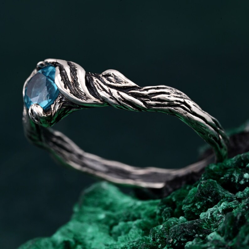 May include: A silver ring with a round, light blue gemstone. The ring's band has a textured, twisted design, resembling a tree branch. The ring is set against a dark green background.