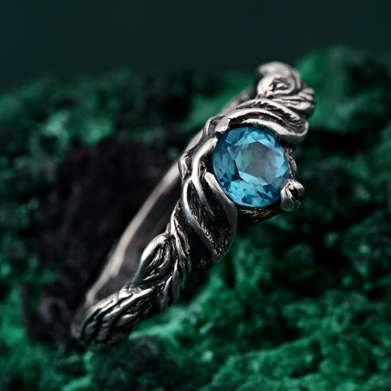 May include: A silver ring with a round, light blue gemstone. The ring has a detailed, nature-inspired design with a textured band and leaf-like accents around the gem. The ring is set against a dark green background.
