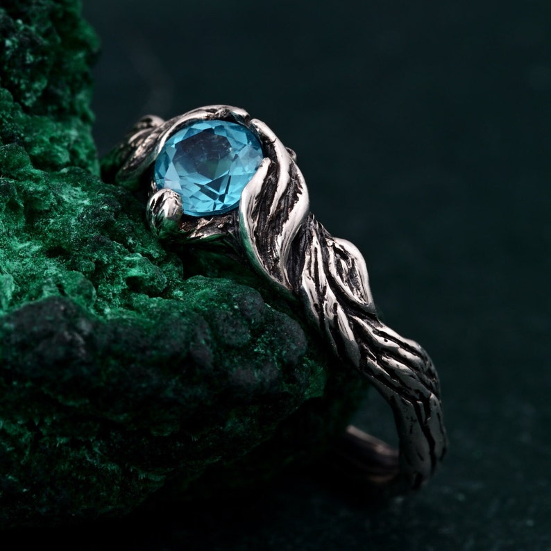 May include: A silver ring with a light blue gemstone. The ring's band and setting have a textured, organic design, resembling intertwined branches. The gemstone is round and sits prominently in the center, creating a striking contrast against the silver.