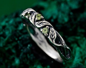 May include: A silver ring with a leafy design and three green gemstones. The ring is on a green background.