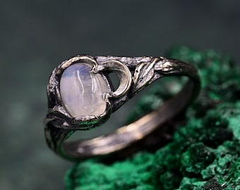 May include: A dark oxidized silver ring featuring an oval-shaped, translucent moonstone cabochon set within a band designed with textured, branch-like details and a small crescent moon accent on one side of the central gemstone setting.