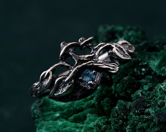 Op de afbeelding: Een zilverkleurige ring met een op de natuur geïnspireerd ontwerp. De ring heeft een gedetailleerd tak- en bladerenmotief, met een kleine, ronde, blauwe edelsteen in het midden. De ring wordt getoond op een getextureerd, donkergroen oppervlak.