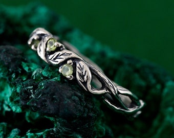 May include: A silver ring featuring a delicate vine design and three green gemstones. The ring is set against a green background.