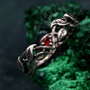 Peut inclure: Une bague argentée avec un motif de feuilles et de vigne. La bague présente une petite pierre précieuse rouge et ronde au centre. Le design de la bague comprend des feuilles détaillées et des vignes torsadées.