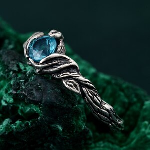 May include: A silver ring with a nature-inspired design. The ring features a large, oval-cut, light blue gemstone held by detailed, textured metalwork resembling intertwined branches. The ring is set against a dark green background.