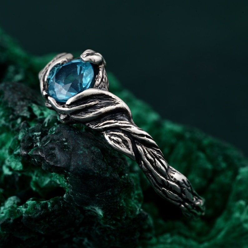 May include: A silver ring with a nature-inspired design. The ring features a large, oval-cut, light blue gemstone held by detailed, textured metalwork resembling intertwined branches. The ring is set against a dark green background.