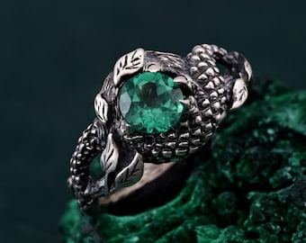 May include: A silver ring with a green gemstone set in a detailed design resembling a dragon or a mythical creature. The ring is set on a green background.