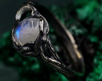 May include: A ring featuring a round, translucent moonstone with a blue iridescent sheen, set within a dark, oxidized metal band that has a textured, branch-like appearance with organic, uneven contours wrapping around the central gemstone.
