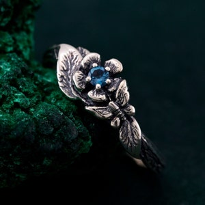 Può includere: Un anello d'argento con un design floreale e una gemma blu al centro. L'anello presenta foglie dettagliate e una farfalla. L'anello è posto su uno sfondo verde scuro.