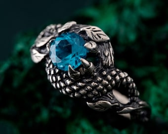 May include: A silver-toned ring featuring a large, round, blue gemstone held in a claw-like setting. The ring has a detailed, textured design with leaf accents, creating a fantasy or gothic aesthetic. The gemstone is a vibrant, eye-catching focal point.