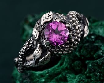 May include: A silver-toned ring featuring a large, round, faceted pink gemstone. The ring's band is intricately designed with a textured, scale-like pattern and leaf accents, creating a nature-inspired aesthetic. The gemstone is the focal point, set prominently in the center.