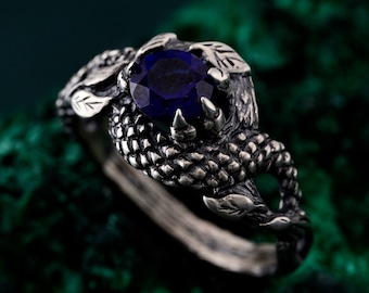 May include: A silver ring featuring a large, oval-cut, deep blue gemstone. The ring's design incorporates intricate details, including claw-like prongs holding the stone and decorative leaf accents. The band has a textured, scale-like pattern.