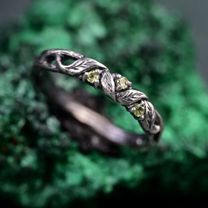 May include: A silver ring with a leaf and vine design. The ring features three small, round, yellow-green gemstones. The band has a textured, organic appearance, with detailed leaf and vine patterns.