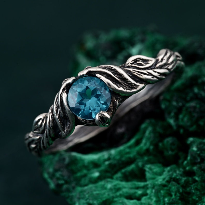 May include: A silver ring with a round, teal-colored gemstone. The ring's band features a textured, organic design, resembling intertwined vines. The gemstone is set in a bezel setting, and the ring is displayed against a green, textured background.