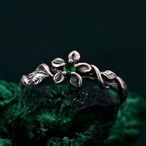 Può includere: Un anello d'argento con una pietra preziosa verde incastonata in un design floreale. L'anello è circondato da foglie e viti.