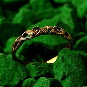 Może przedstawiać: Złoty pierścionek o wzorze inspirowanym naturą, z trzema ciemnymi kamieniami szlachetnymi. Pierścionek jest osadzony na tle zielonych kamieni, podkreślając jego skomplikowane detale i elegancki styl. Projekt pierścionka obejmuje motywy liści i gałęzi.