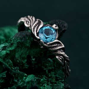 May include: A silver ring featuring a large, round, light blue gemstone. The ring's band has a detailed, leaf-like design that wraps around the gemstone. The ring is set against a dark green, textured background.