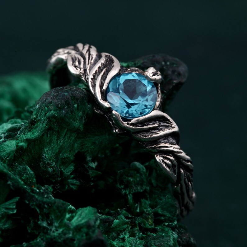 May include: A silver ring featuring a large, round, light blue gemstone. The ring's band has a detailed, leaf-like design that wraps around the gemstone. The ring is set against a dark green, textured background.