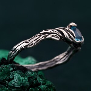 May include: A silver-toned ring with a branch-like design. The ring features a teardrop-shaped, light blue gemstone. The band has a textured, organic appearance. The ring is set against a dark green background.