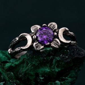 Könnte beinhalten: Ein silberner Ring mit einem violetten Edelstein, der in einem Blumenmuster gefasst ist. Der Ring hat außerdem zwei Halbmondformen auf beiden Seiten der Blume.