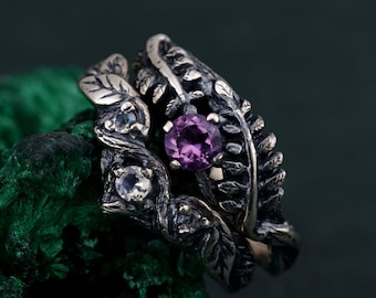 May include: Two silver rings with leaf and vine detailing. One ring features a large, round, purple gemstone, while the other has smaller, clear gemstones. The rings are set against a dark green background.