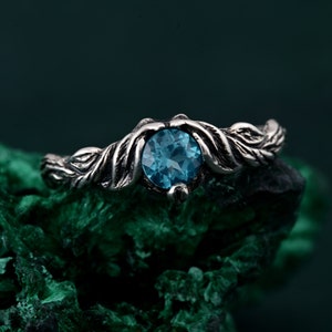 May include: A silver ring with a round, light blue gemstone. The ring's band features a detailed leaf design. The gemstone is set in a decorative setting, creating a nature-inspired aesthetic. The ring is displayed on a textured, dark green surface.