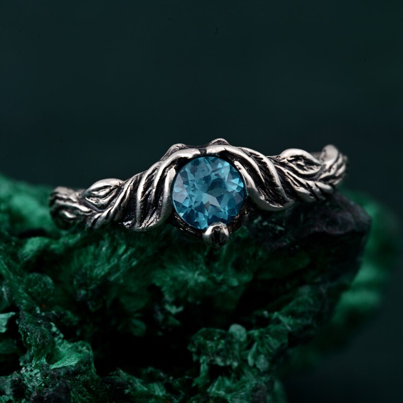 May include: A silver ring with a round, light blue gemstone. The ring's band features a detailed leaf design. The gemstone is set in a decorative setting, creating a nature-inspired aesthetic. The ring is displayed on a textured, dark green surface.