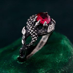 Puede incluir: Un anillo de plata con una piedra preciosa roja engastada en una garra. El anillo está diseñado para parecer una garra de dragón.